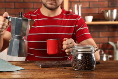 Brewing coffee. Man with jar of beans, moka pot and mug at wooden table indoors, closeup