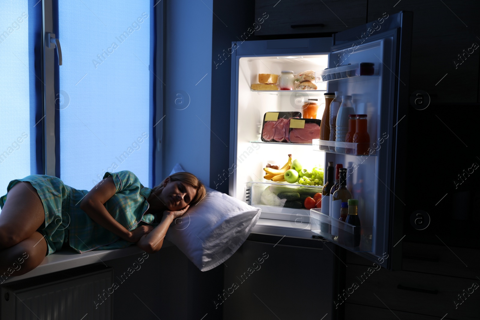Photo of Woman sleeping on window sill near open refrigerator in kitchen at night