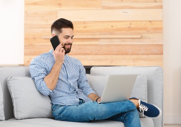 Photo of Handsome young man talking on phone while working with laptop on sofa at home