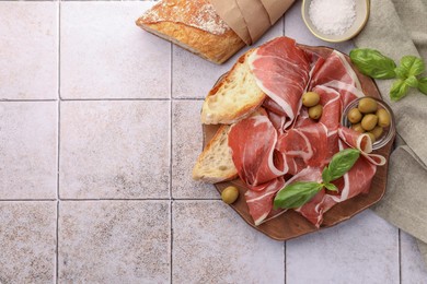 Photo of Slices of tasty cured ham, olives, bread and basil on tiled table, flat lay. Space for text