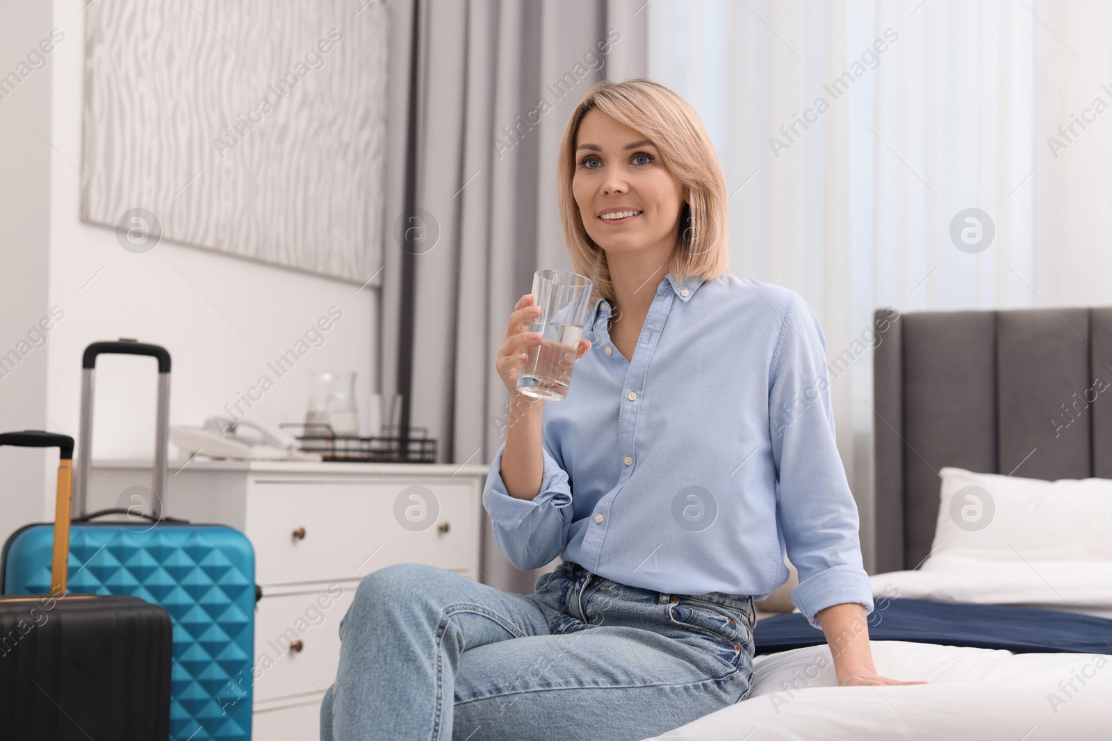 Photo of Smiling guest with glass of water on bed in stylish hotel room