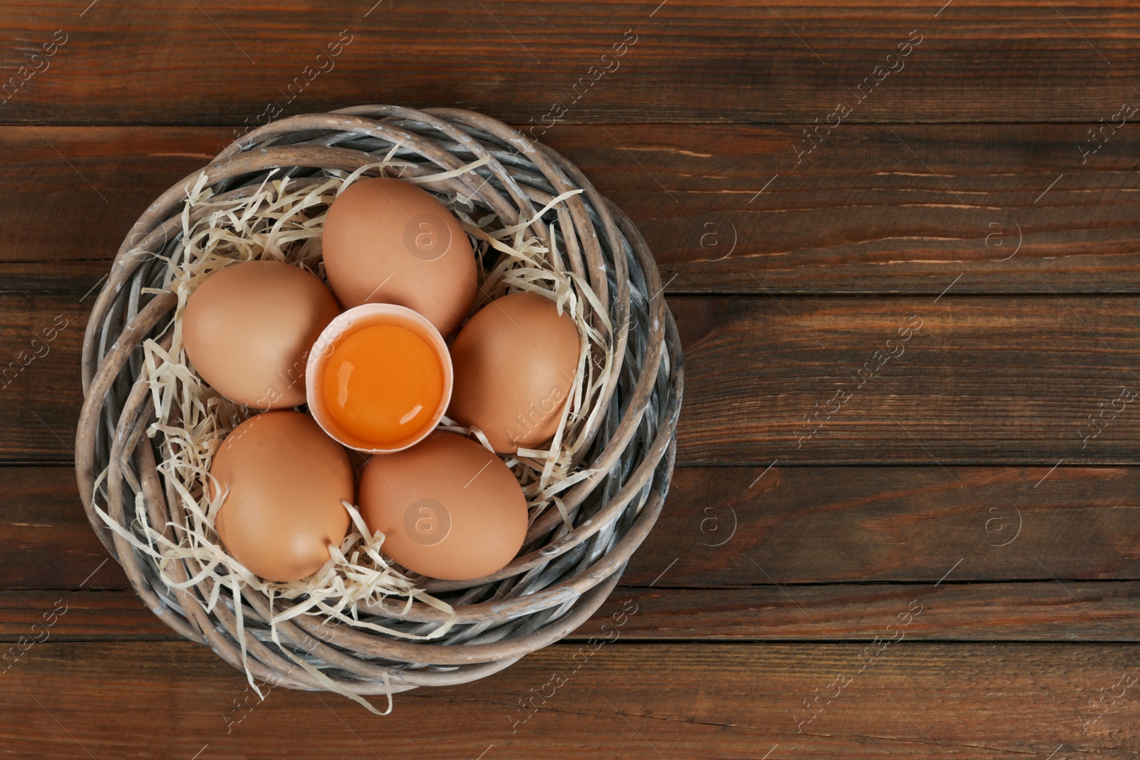 Photo of Raw chicken eggs in wicker nest on wooden table, top view