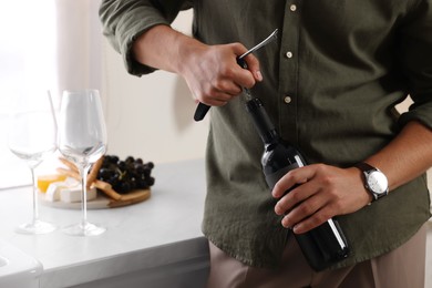 Photo of Romantic dinner. Man opening wine bottle with corkscrew in kitchen, closeup