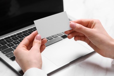 Photo of Woman with laptop holding blank business card at white table, closeup. Space for text