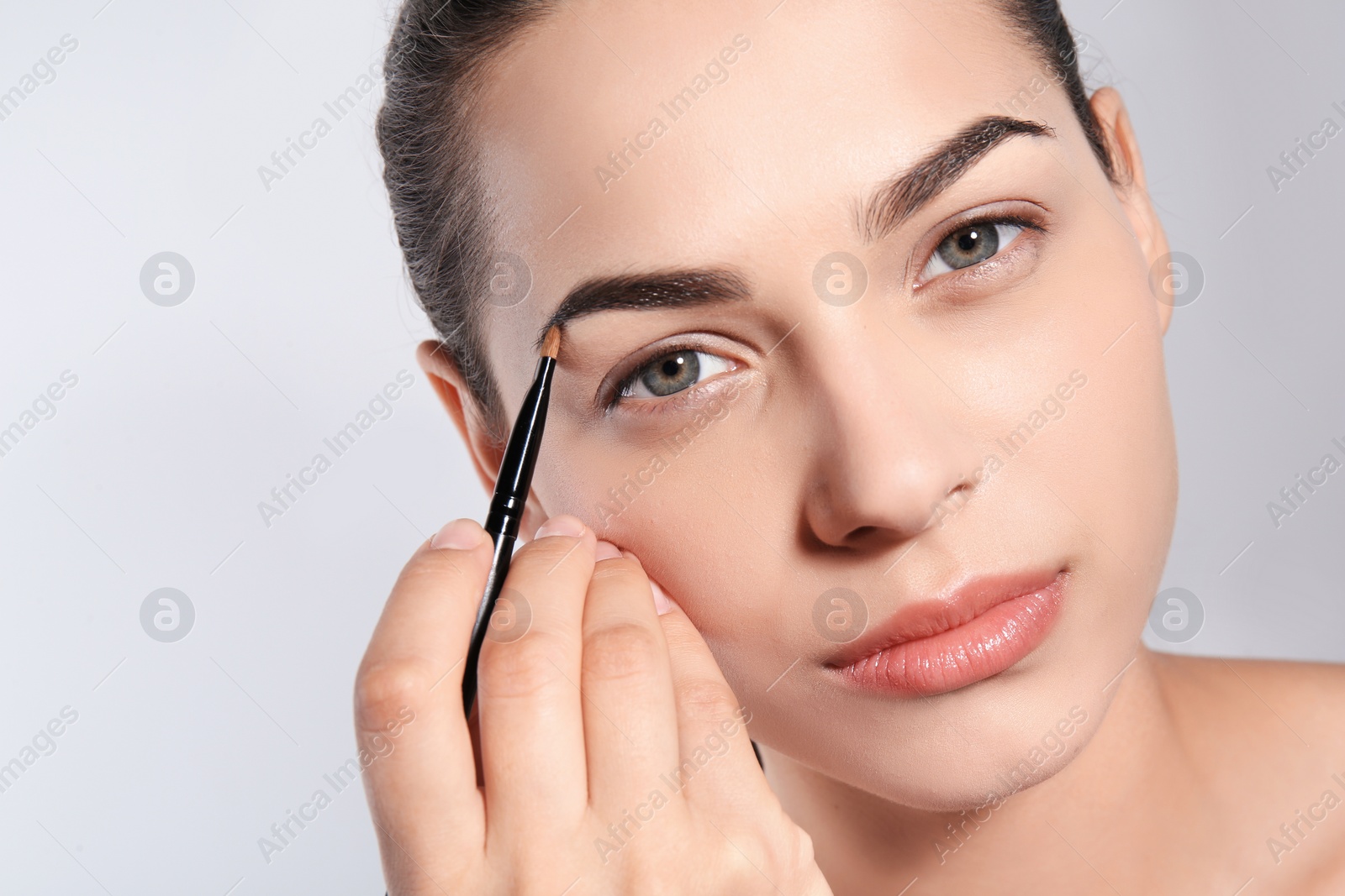 Photo of Young woman correcting shape of eyebrow with brush on light background