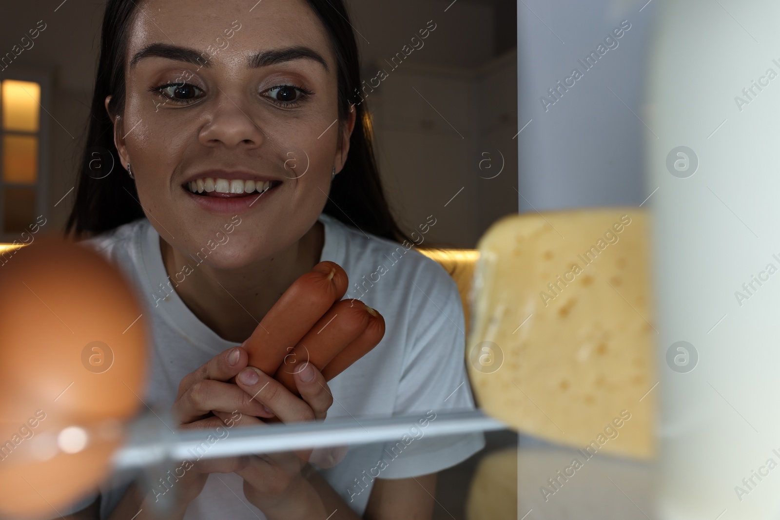 Photo of Young woman taking sausages out of refrigerator in kitchen at night, view from inside