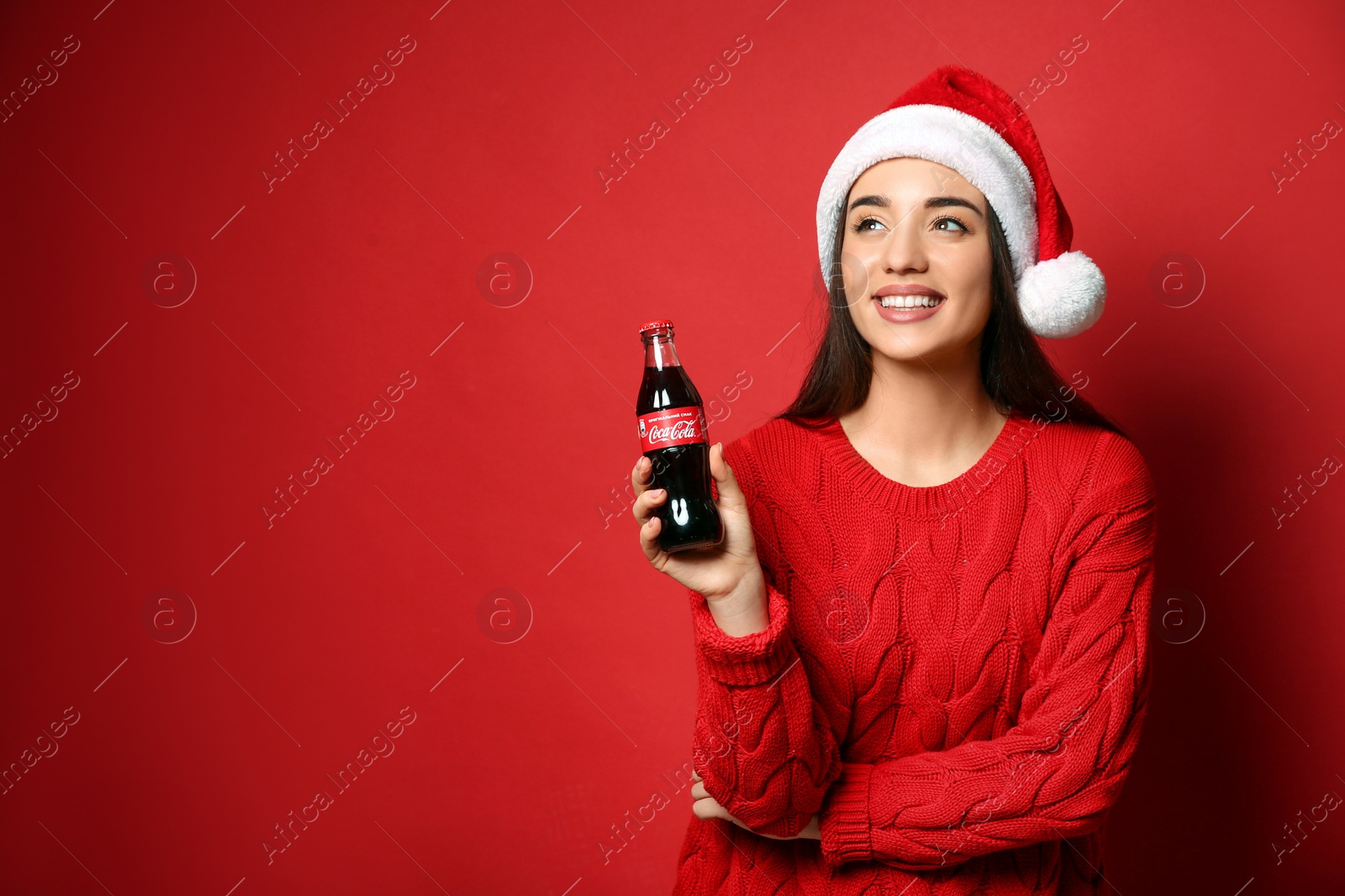 Photo of MYKOLAIV, UKRAINE - JANUARY 27, 2021: Young woman in Christmas hat holding bottle of Coca-Cola on red background. Space for text