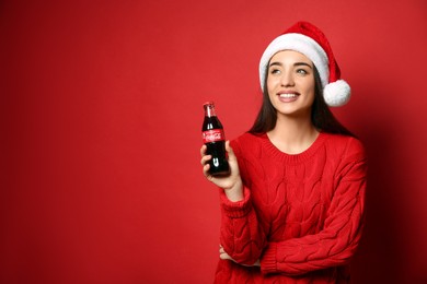 Photo of MYKOLAIV, UKRAINE - JANUARY 27, 2021: Young woman in Christmas hat holding bottle of Coca-Cola on red background. Space for text
