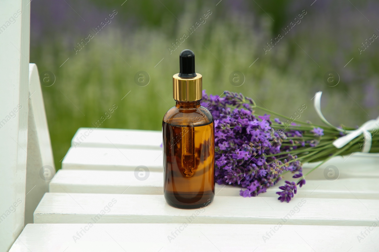 Photo of Bottle of essential oil and lavender flowers on white wooden table in field