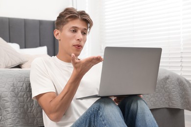 Photo of Happy young man having video chat via laptop and blowing kiss indoors. Long-distance relationship