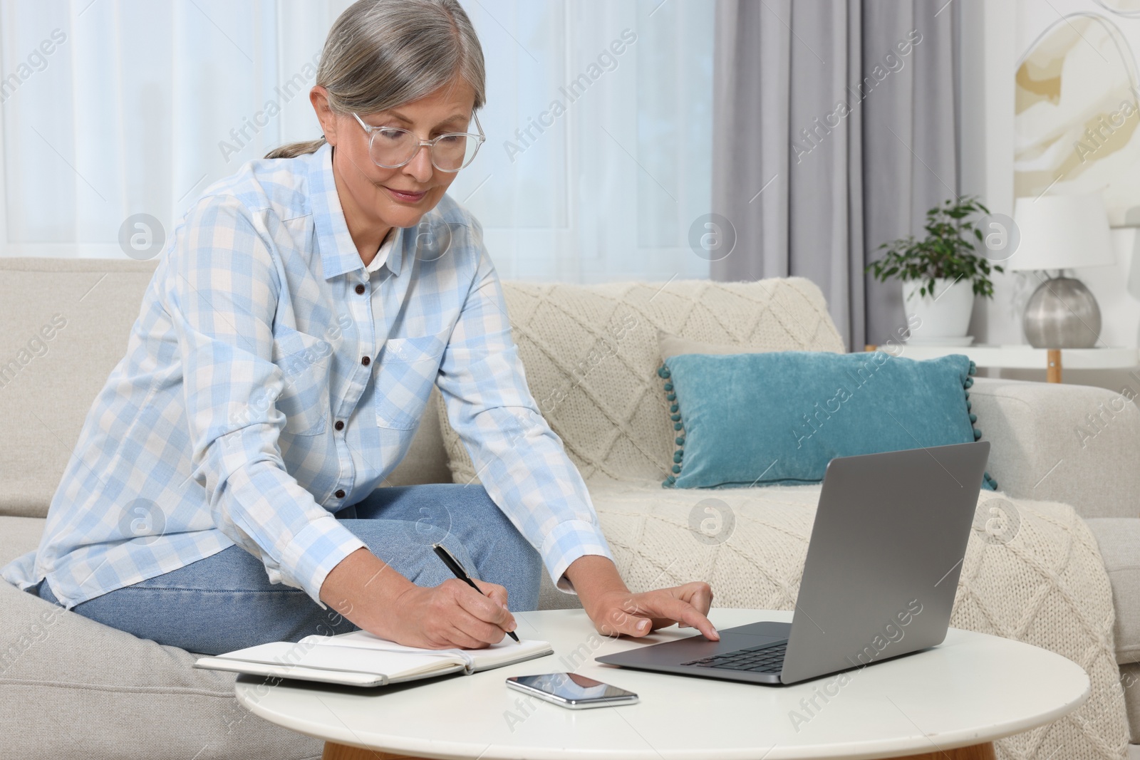 Photo of Beautiful senior woman writing something in notebook while using laptop at home