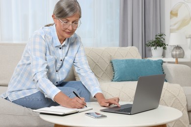 Photo of Beautiful senior woman writing something in notebook while using laptop at home