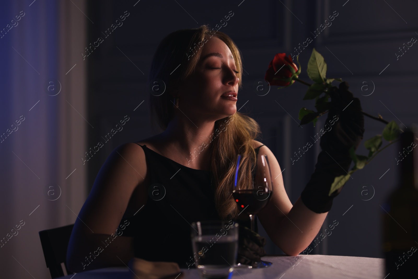 Photo of Elegant young woman with glass of wine and rose at table indoors in evening