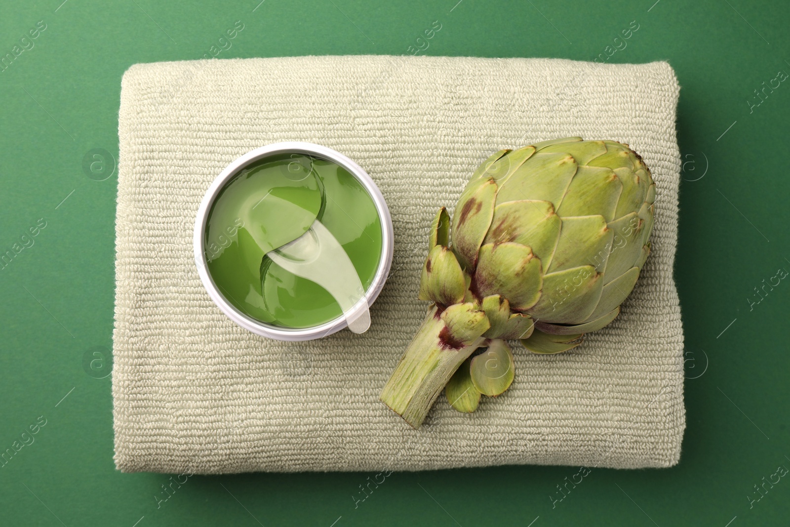 Photo of Package of under eye patches, towel and artichoke on green background, top view. Cosmetic product