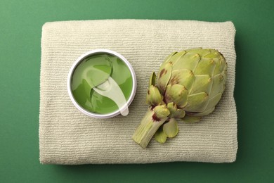 Photo of Package of under eye patches, towel and artichoke on green background, top view. Cosmetic product