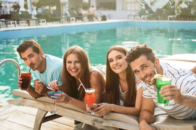 Happy young friends with fresh summer cocktails relaxing near swimming pool