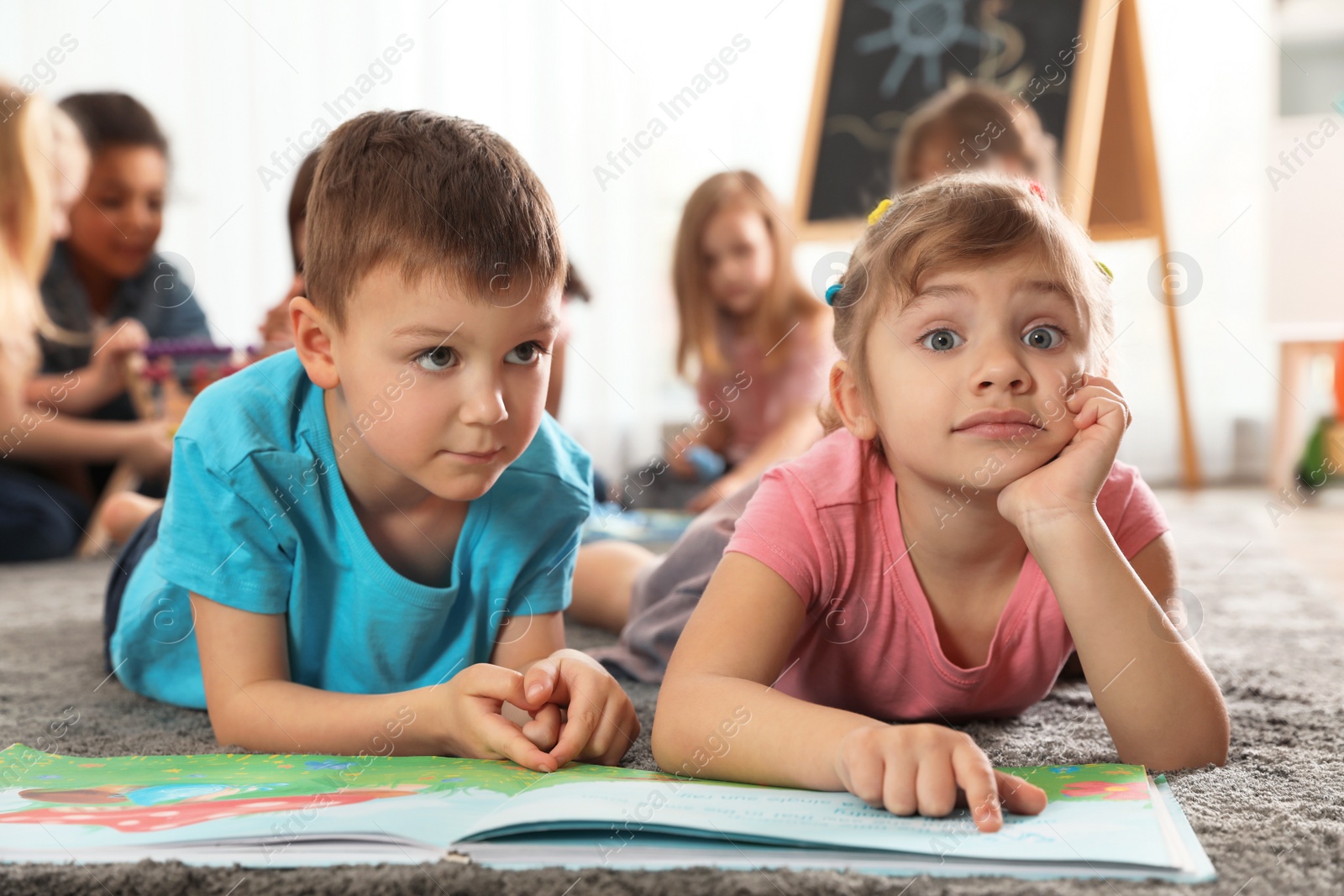 Photo of Cute kids reading book on floor while other children playing together in kindergarten