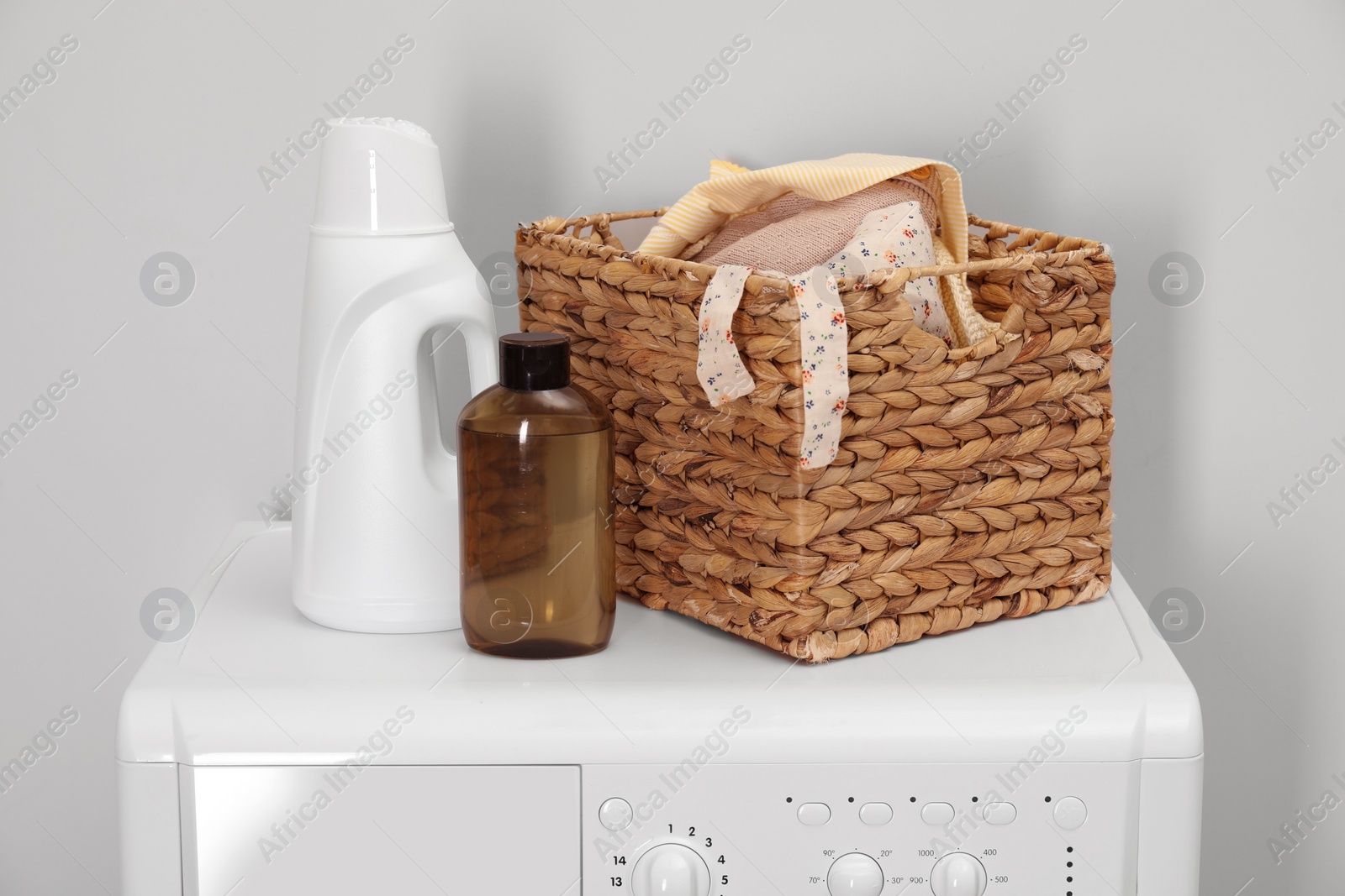 Photo of Baby clothes in wicker basket and laundry detergents on washing machine near light wall