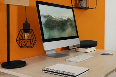 Photo of Modern computer, books, lamp and notebook on wooden desk near orange wall. Home office