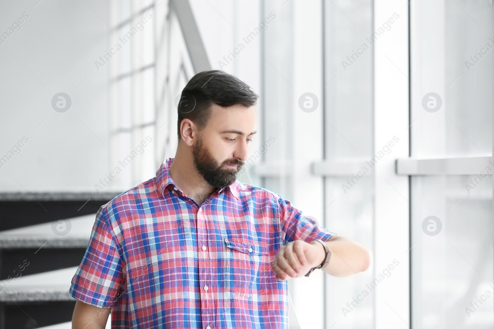 Photo of Portrait of handsome bearded man looking at wristwatch near stairs, indoors