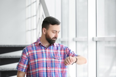 Portrait of handsome bearded man looking at wristwatch near stairs, indoors