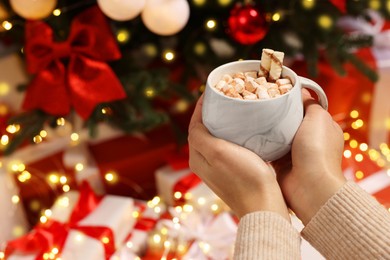 Photo of Woman holding cup of delicious cocoa with marshmallows and wafer sticks near Christmas tree and gifts, closeup. Space for text