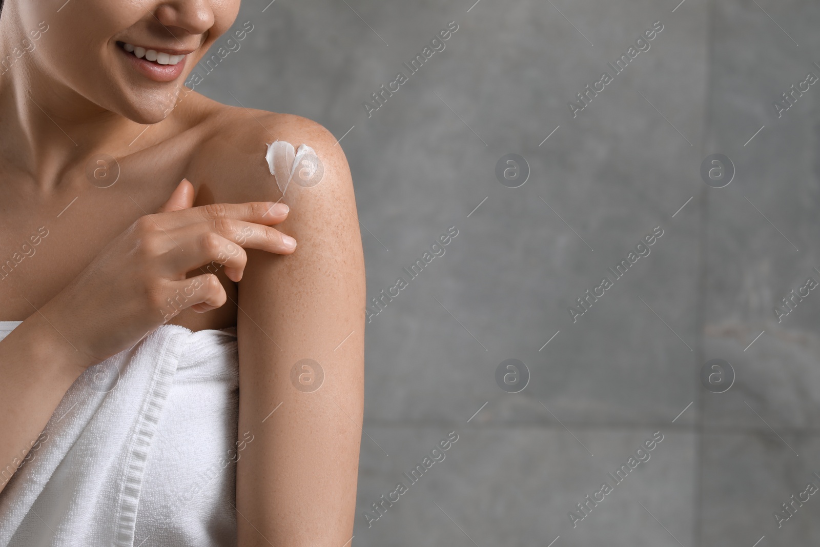 Photo of Young woman applying body cream onto shoulder near grey wall, closeup. Space for text