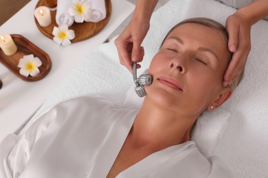 Photo of Woman receiving facial massage with metal roller in beauty salon, above view