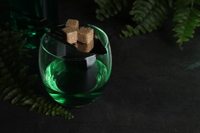 Photo of Absinthe in glass, spoon, brown sugar cubes and fern leaves on gray textured table, closeup with space for text. Alcoholic drink