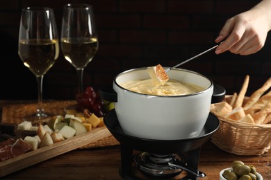 Photo of Woman dipping piece of bread into fondue pot with melted cheese at wooden table, closeup