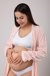 Photo of Beautiful pregnant woman in stylish comfortable underwear and robe making heart with hands on her belly against grey background