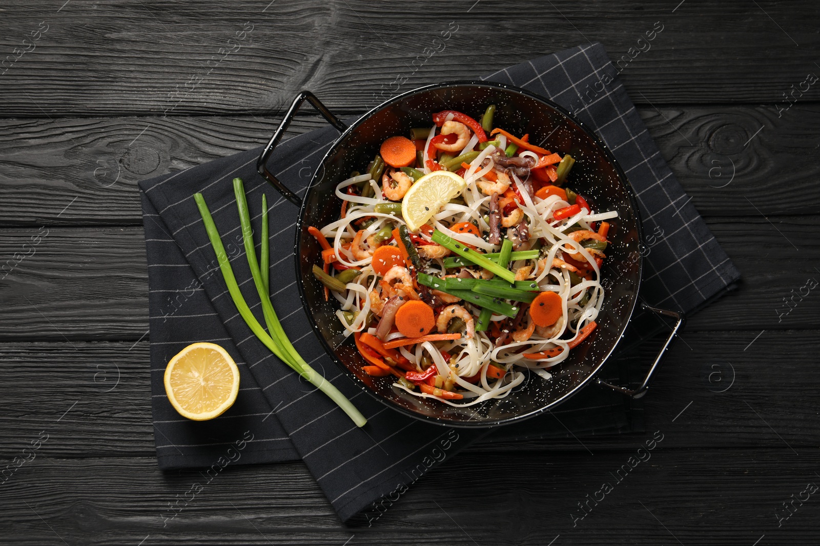 Photo of Shrimp stir fry with noodles and vegetables in wok on black wooden table, top view