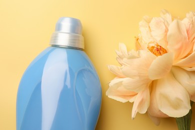 Photo of Bottle of fabric softener and beautiful peony flower on pale yellow background, flat lay