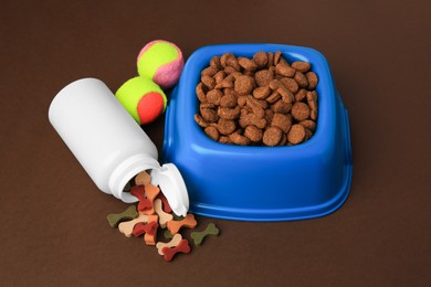 Bowl with dry pet food, bottle of vitamins and toy balls on brown background