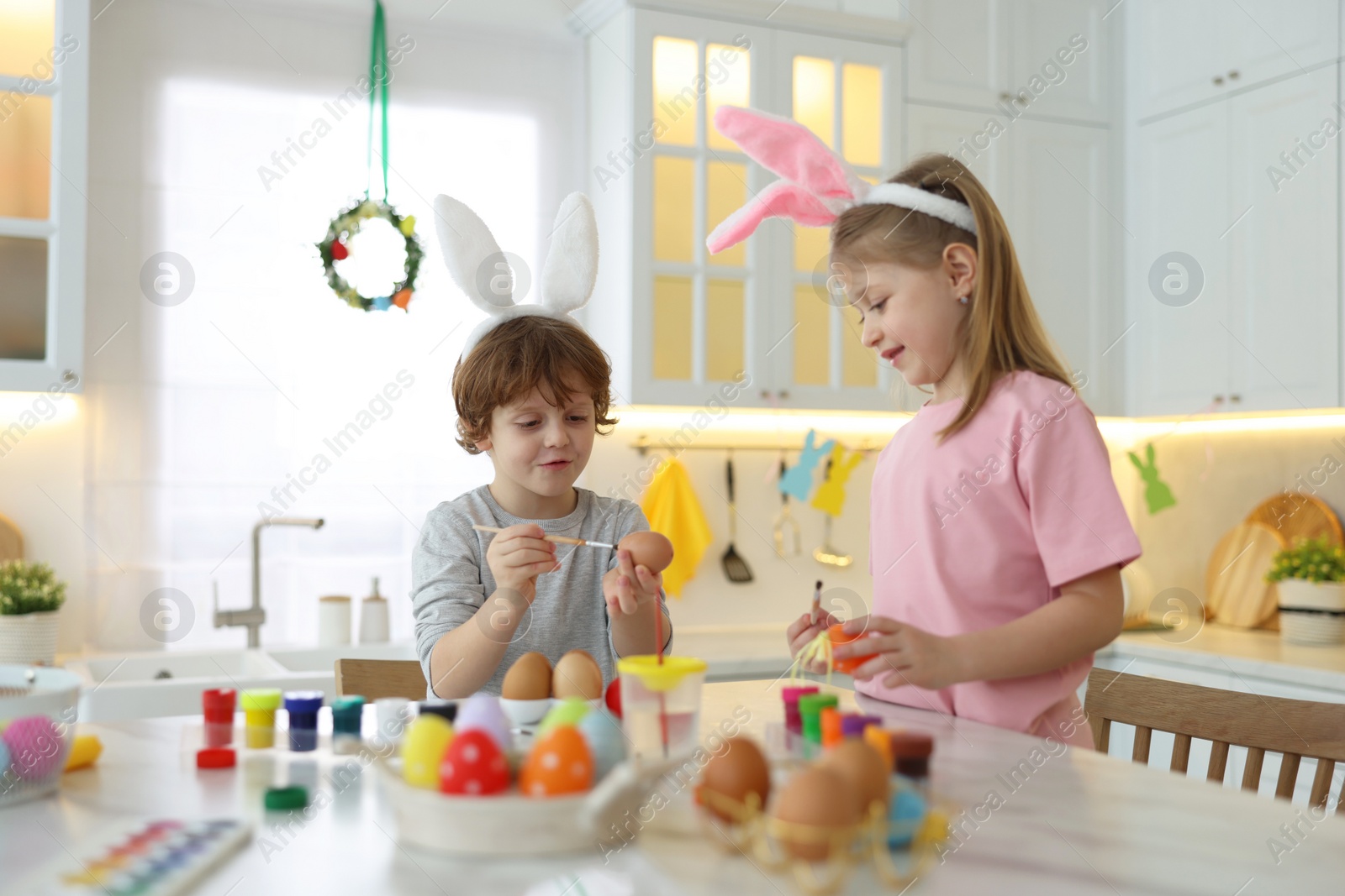 Photo of Easter celebration. Cute children with bunny ears painting eggs at white marble table in kitchen