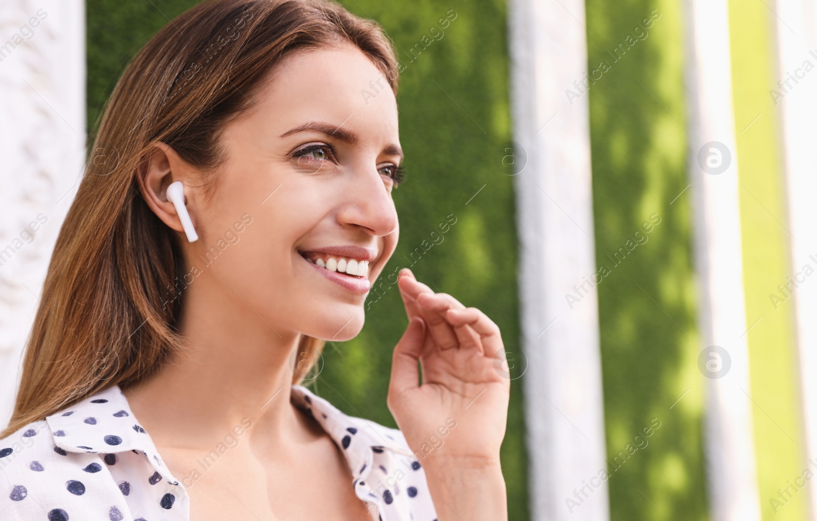 Photo of Young woman with wireless headphones listening to music near green grass wall. Space for text
