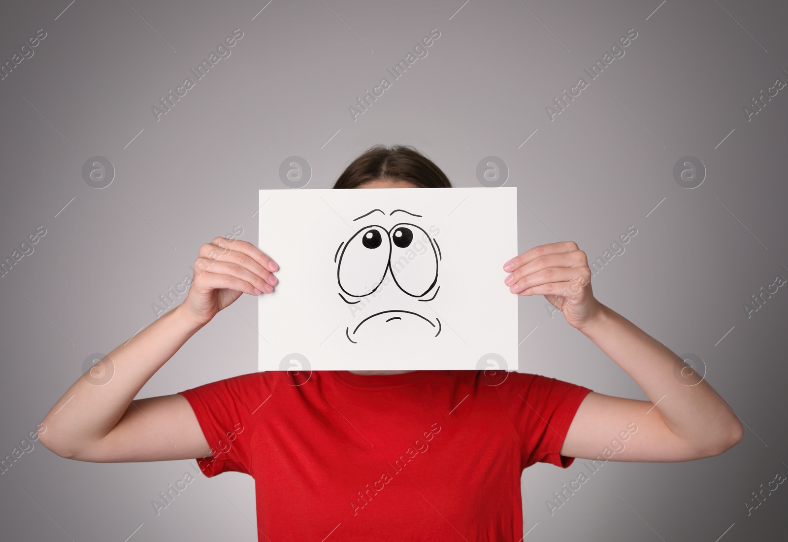 Photo of Woman hiding behind sheet of paper with anxious face on grey background