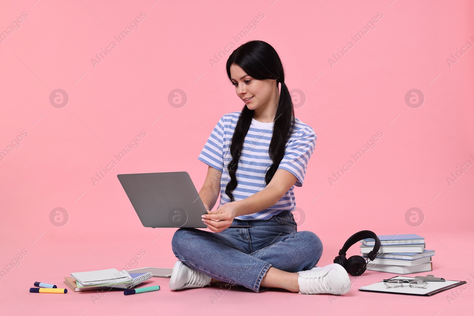Photo of Student with laptop sitting among books and stationery on pink background