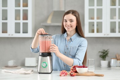 Beautiful young woman preparing tasty smoothie at white table in kitchen