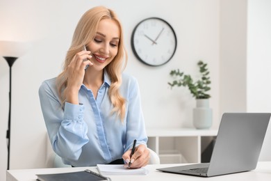 Photo of Happy secretary taking notes while talking on smartphone in office