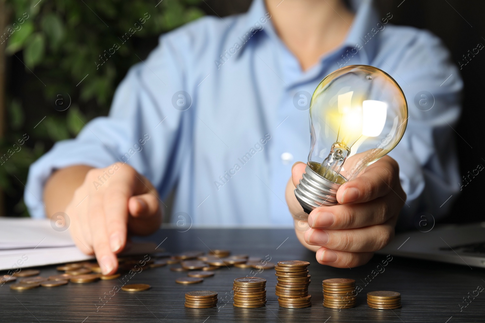 Photo of Woman with light bulb and coins at black wooden table, closeup. Power saving