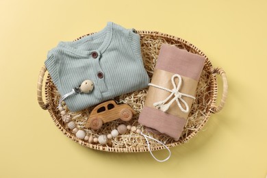 Different baby accessories and clothes in wicker basket on yellow background, top view