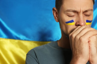 Sad man with clasped hands near Ukrainian flag, closeup. Space for text