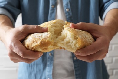 Photo of Man breaking loaf of fresh bread near white brick wall, closeup