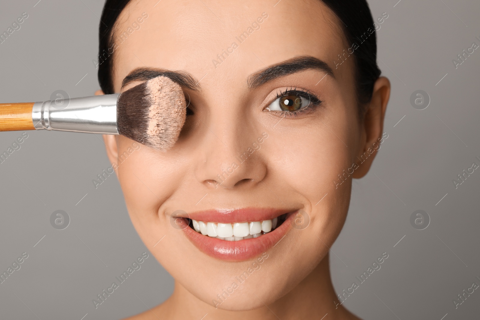 Photo of Beautiful young woman with face powder on brush against grey background, closeup