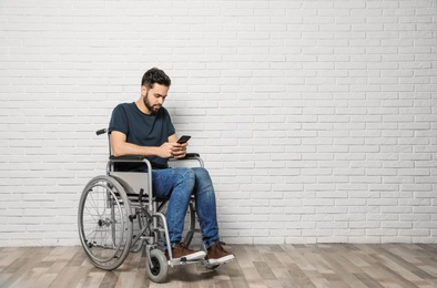 Young man with smartphone in wheelchair near brick wall indoors. Space for text