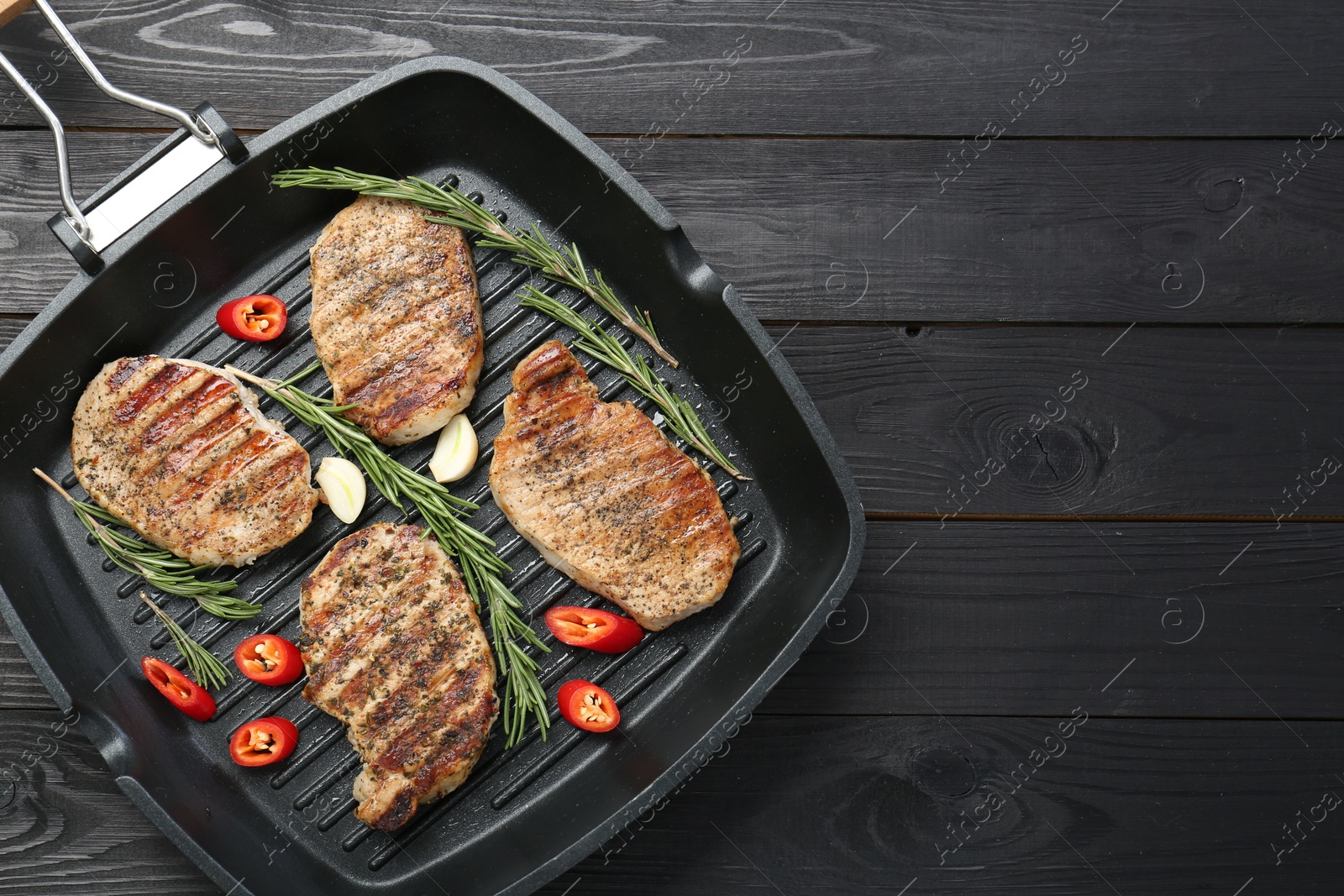 Photo of Grill pan with delicious pork steaks, garlic, chili pepper and rosemary on black wooden table, top view. Space for text