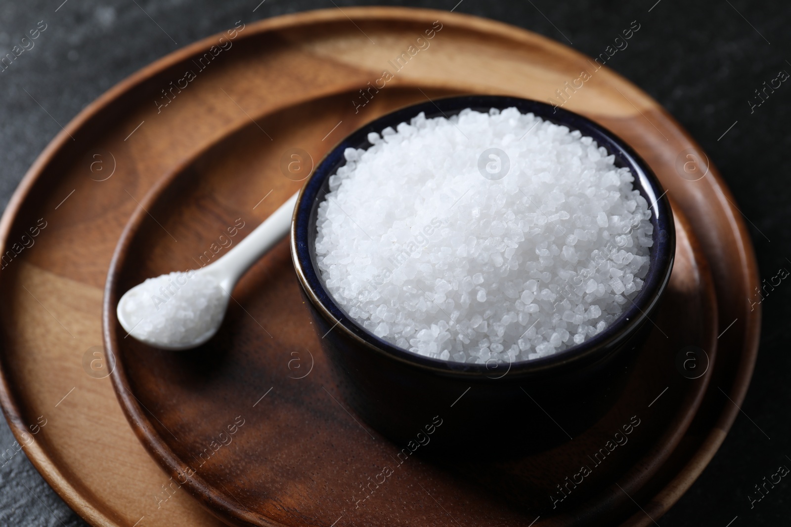 Photo of Organic white salt in bowl and spoon on black table, closeup