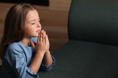 Cute little girl with hands clasped together praying indoors. Space for text