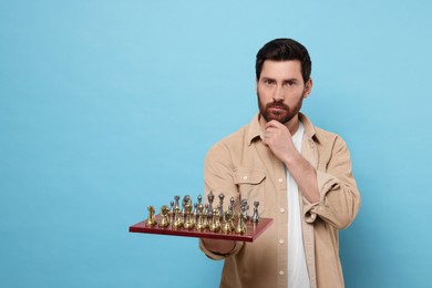 Photo of Thoughtful man holding chessboard with game pieces on light blue background. Space for text